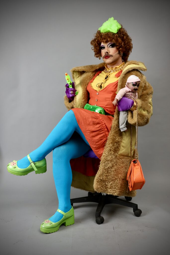 Photo de Lee Shulman de la drag queen Victoria Sucrette, Diabétique vénère. Elle est assise sur une chaise avec des vêtements colorés et porte dans ses mains son bébé qui s'appelle Stévia